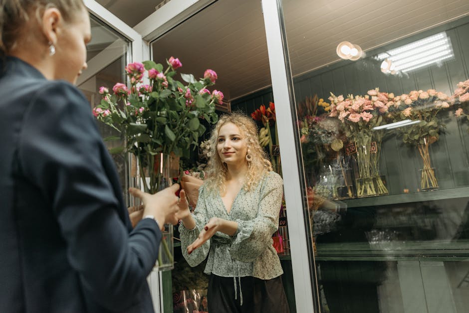 A young woman with curly blonde hair, wearing a light patterned blouse, is inside a flower shop selecting a bouquet of fresh pink roses with green foliage. The bouquet is being handed over by a person