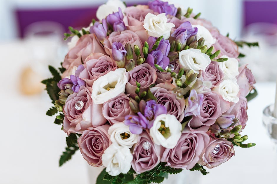 A close-up of a floral bouquet arrangement featuring soft pink roses, white lisianthus, and purple freesia, accented with green foliage. The flowers are freshly cut with vibrant petals and sturdy stem