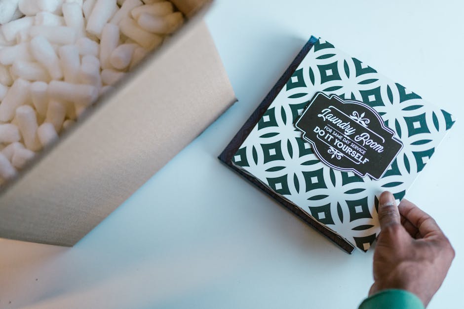 A hand pulls open a decorative gift box with a black and white geometric pattern, revealing white packing peanuts inside. The box, branded with a label that reads 'Laundry Room for same day service, d