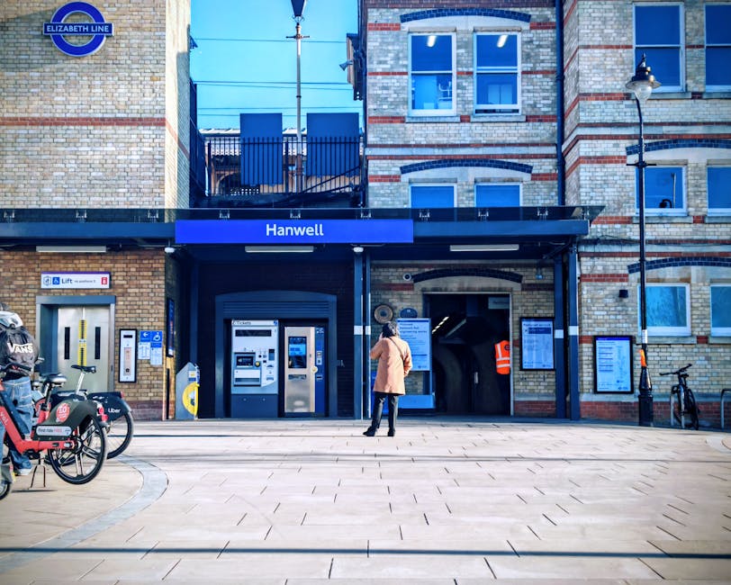 A woman in a beige coat and black hat stands outside Hanwell station, which features a brick facade with a blue sign reading 'Hanwell' above the entrance. The station entrance has automated ticket mac