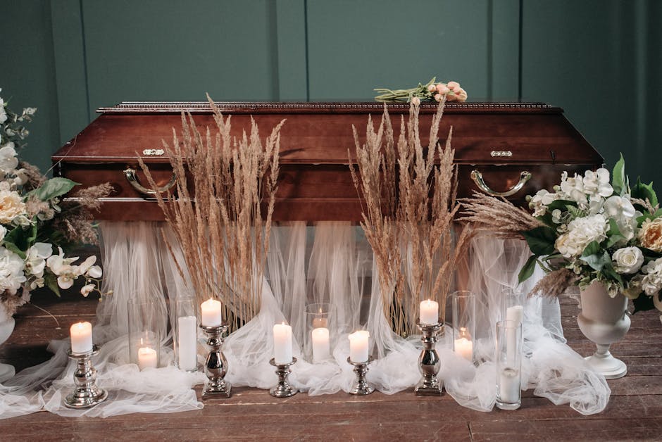 A wooden funeral casket surrounded by floral arrangements and candles in a respectful setting. To the left and right of the casket are large white and cream-colored flowers in vases, including roses a