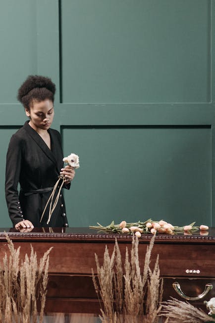 A young woman with dark, curly hair styled in an updo, dressed in a black blazer with gold button details, standing beside a polished wooden casket adorned with a floral arrangement. She holds a small